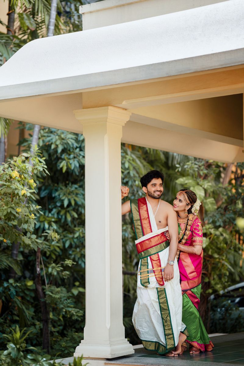 Couple in traditional South Indian attire sharing a playful moment under white pillared gazebo.