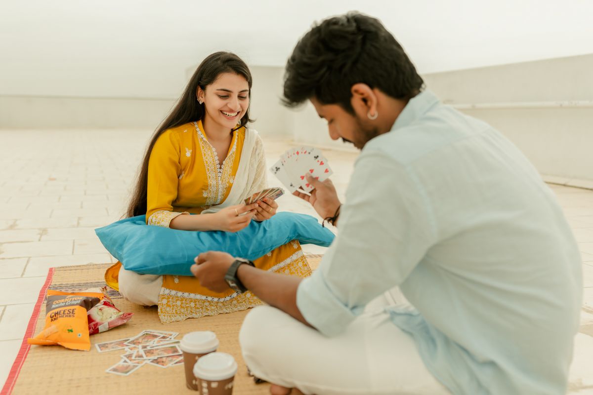 A couple sits on a white floor playing cards together while enjoying snacks.