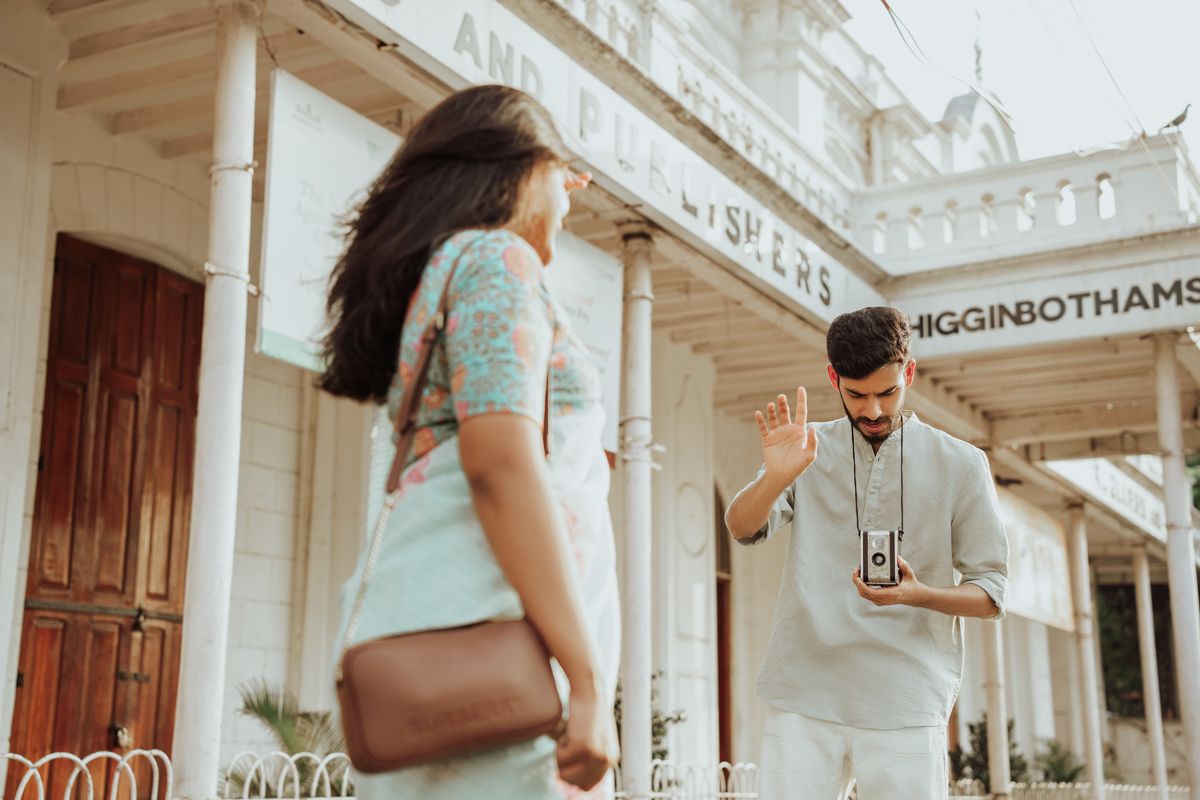 Two people in casual clothing interact on a sunny day in front of a white colonial-style building facade.