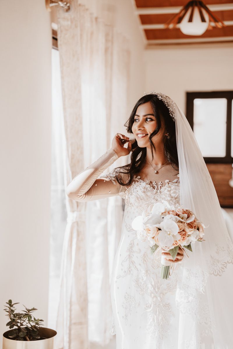 A bride in an elegant lace wedding dress holding a pink and white bouquet while talking on the phone by a window.