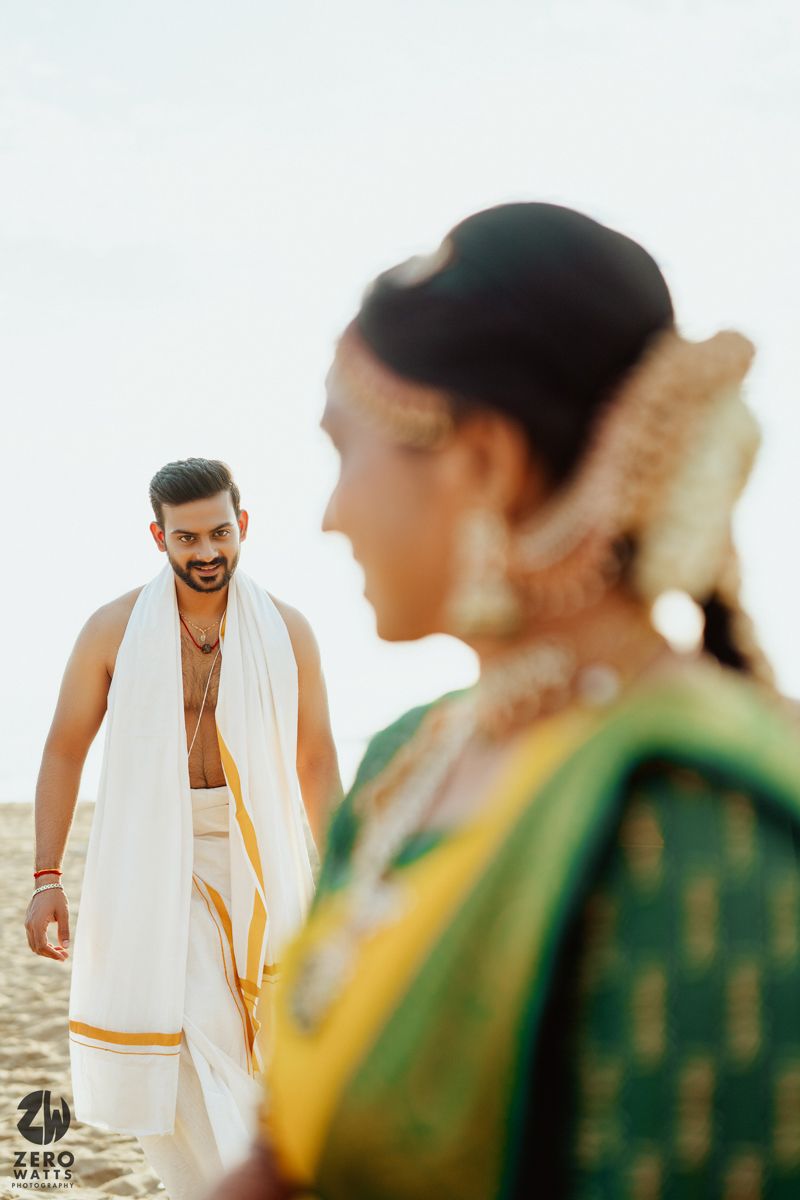 A couple in traditional South Indian attire pose on a beach during sunset with the woman in focus in the foreground.