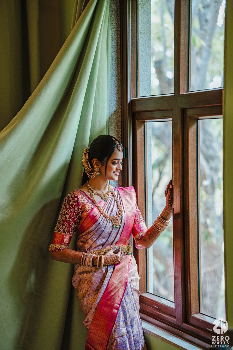 Traditional bride in pink silk saree poses elegantly by window with green curtains.