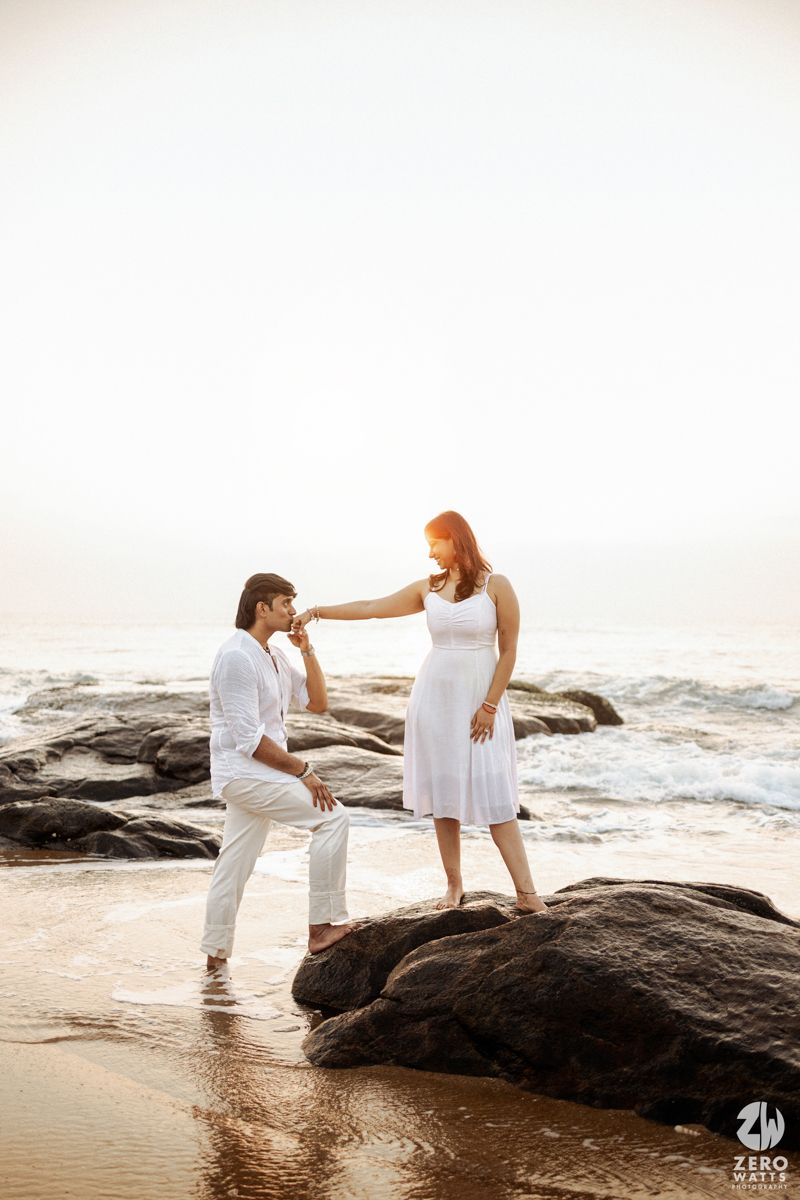 A couple shares a romantic moment on coastal rocks at sunset as waves crash behind them.