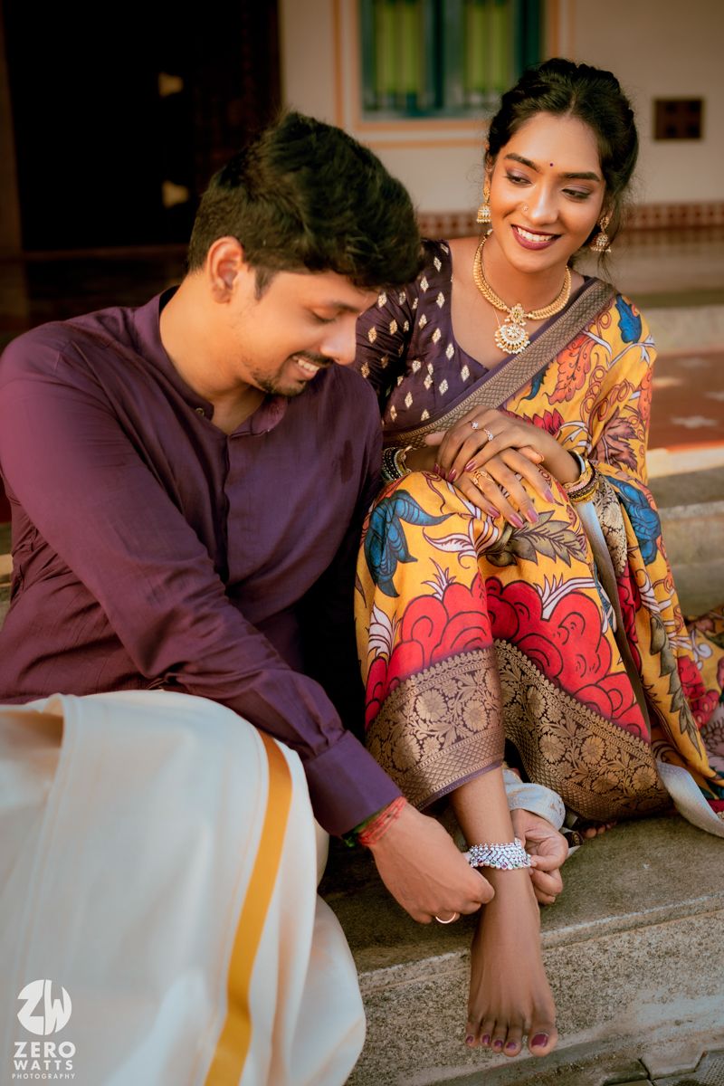 Couple shares intimate moment during traditional Indian toe ring ceremony wearing colorful attire.