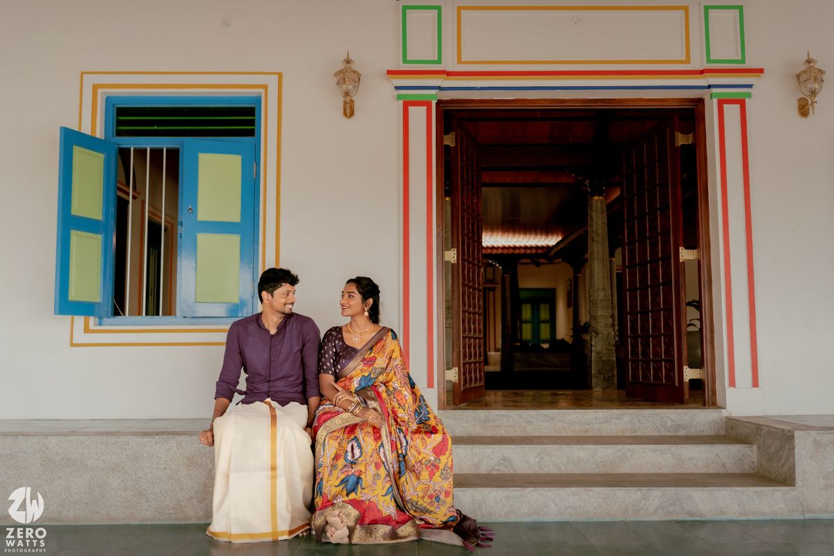 A couple in colorful traditional attire sit together outside a building with blue-framed windows and ornate doorway.