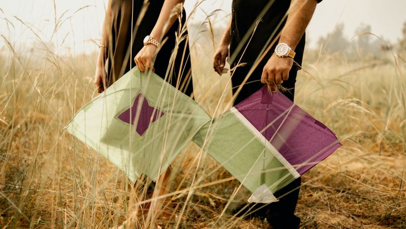 Hands holding green and purple kites in a golden wheat field during sunset.