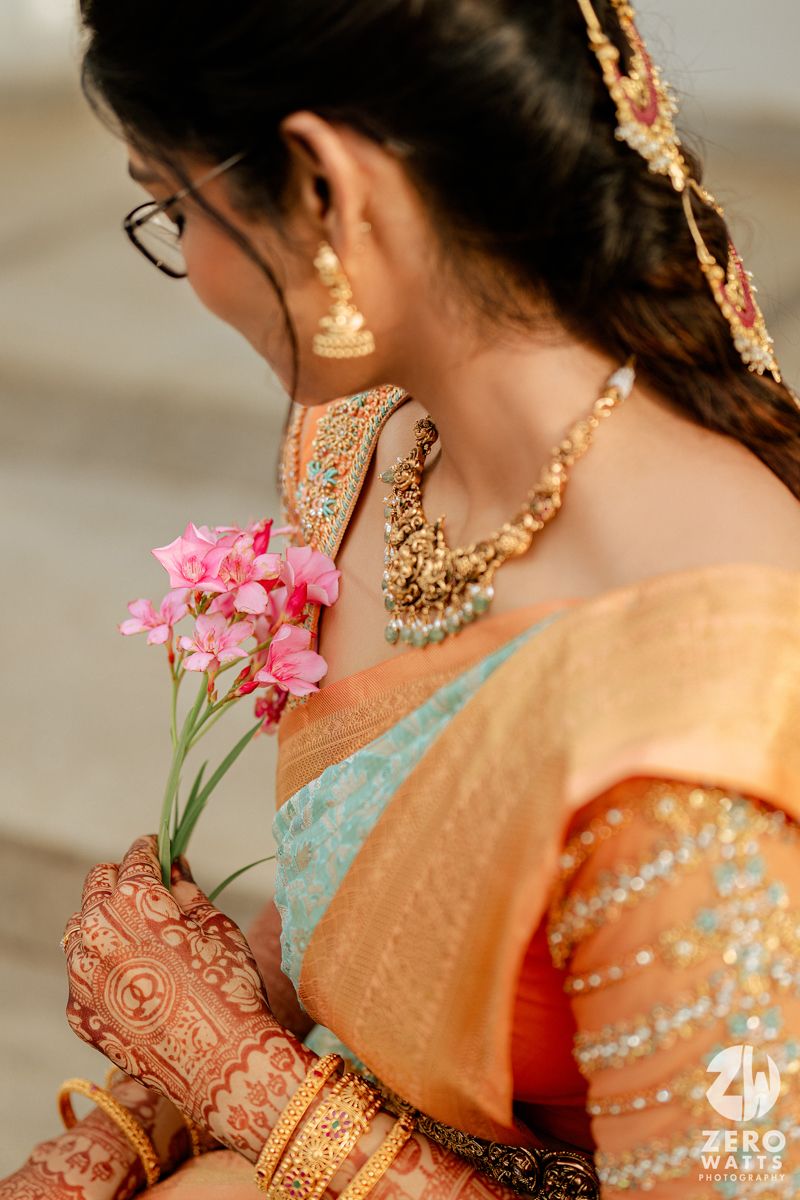Traditional Indian bridal jewelry and mehndi decorated hands holding pink flowers against a peach and mint colored sari.