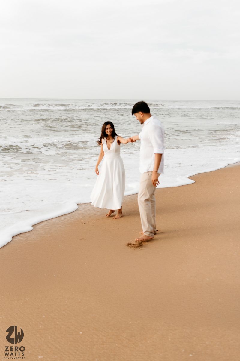 Two people in white clothing walk along a sandy beach with ocean waves rolling in.