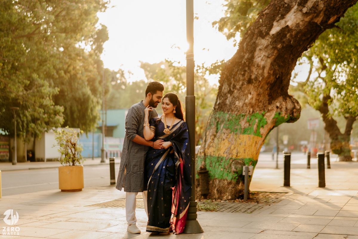 A romantic couple in traditional Indian attire embraces beneath a large tree during golden hour on a city street.