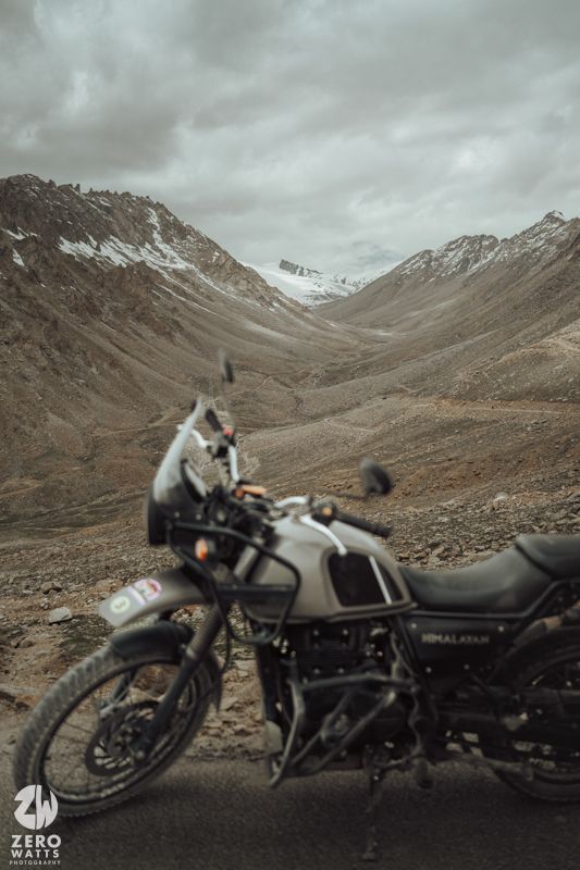 Classic motorcycle parked in rugged mountain landscape with snow-capped peaks under moody overcast skies.