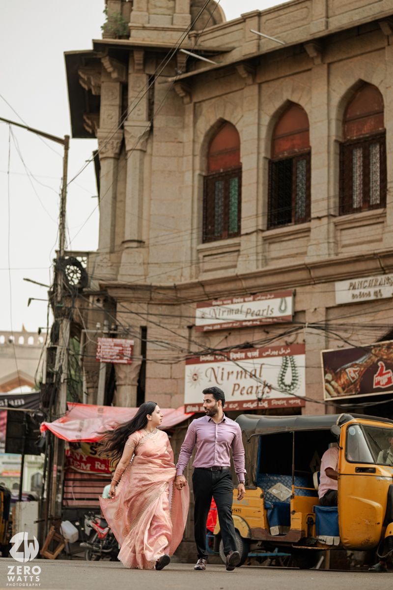 A couple walking through a bustling street in Old City Hyderabad with auto rickshaws and shops in background.