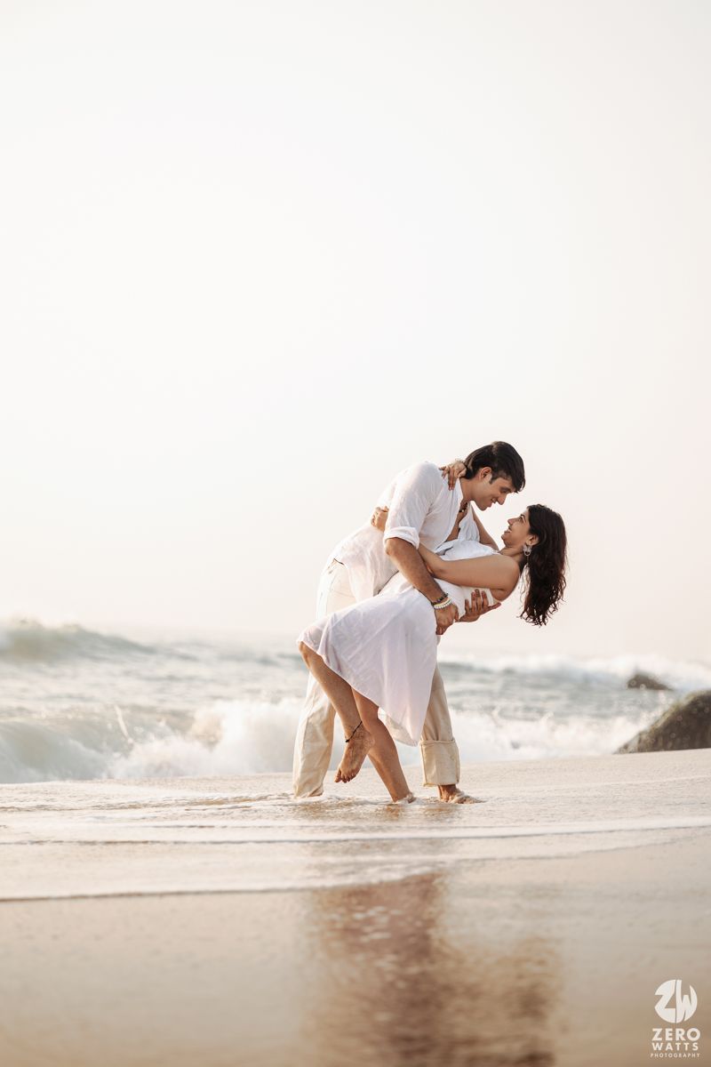 Couple in white clothing dances together on a sandy beach with waves crashing behind them.
