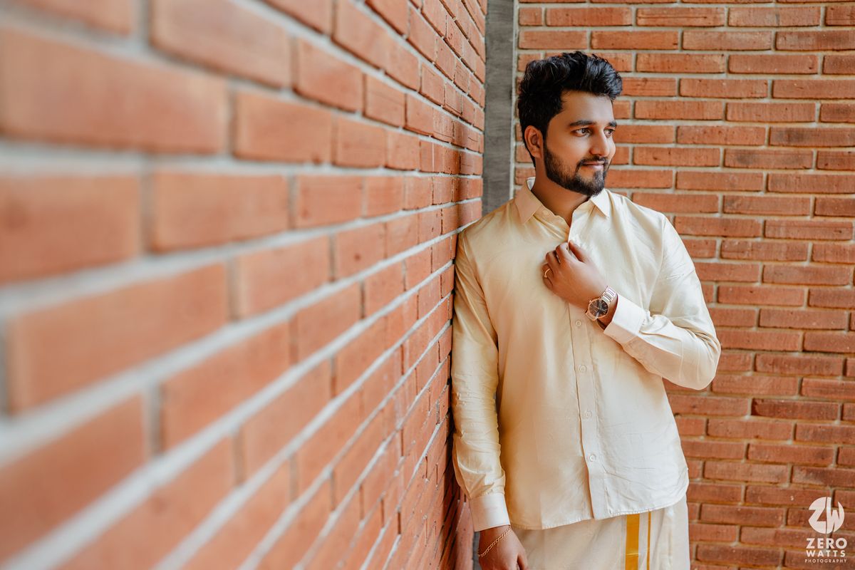 Groom in cream colored traditional Indian formal wear poses against brick wall.