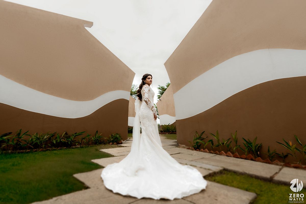 A bride in a flowing white lace gown with long train poses along a curved path between modern architectural walls.