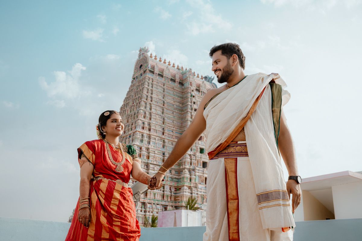 Two people in traditional Indian attire hold hands while standing on a rooftop with a temple tower visible in background.