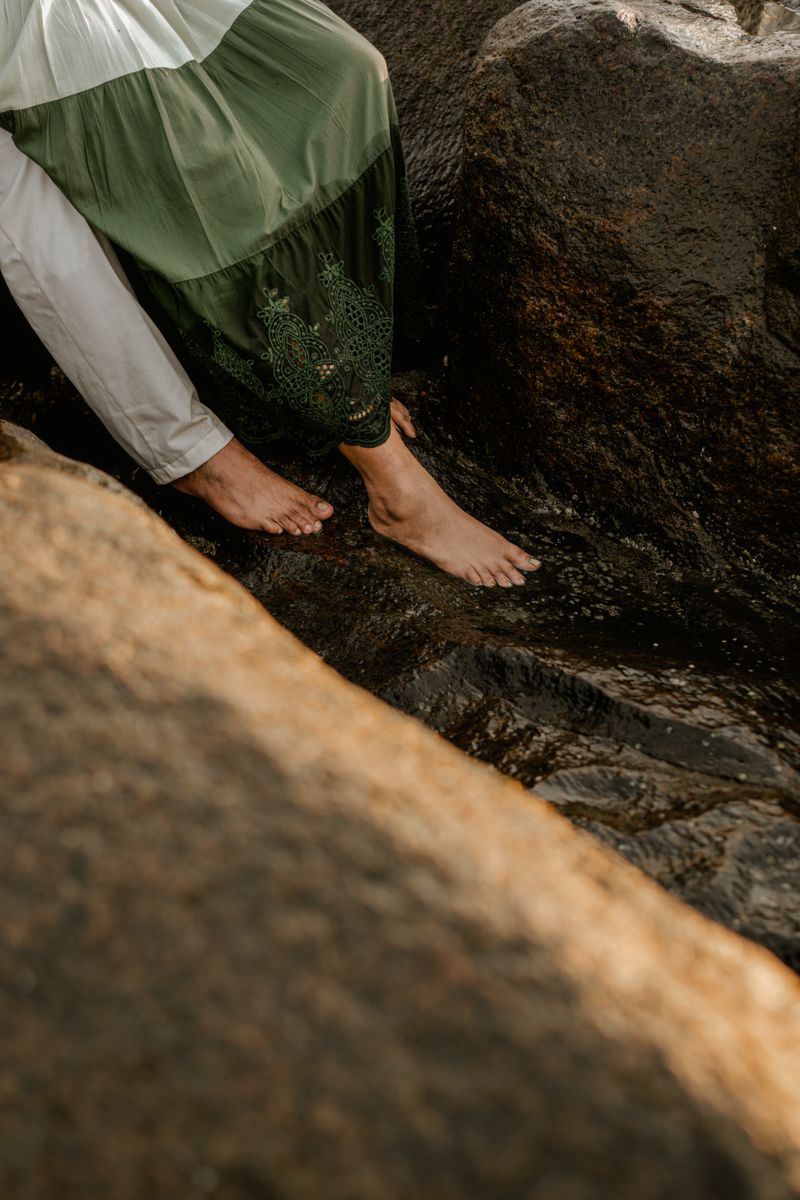Bare feet resting on dark rocks near water, wearing flowy green and white clothing.