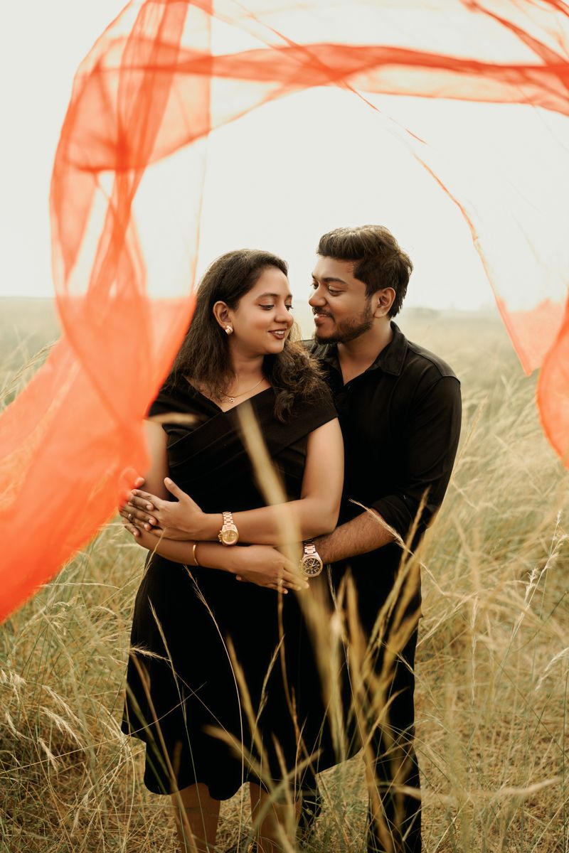 A couple embraces in a field of tall grass while a flowing orange fabric dances in the breeze.