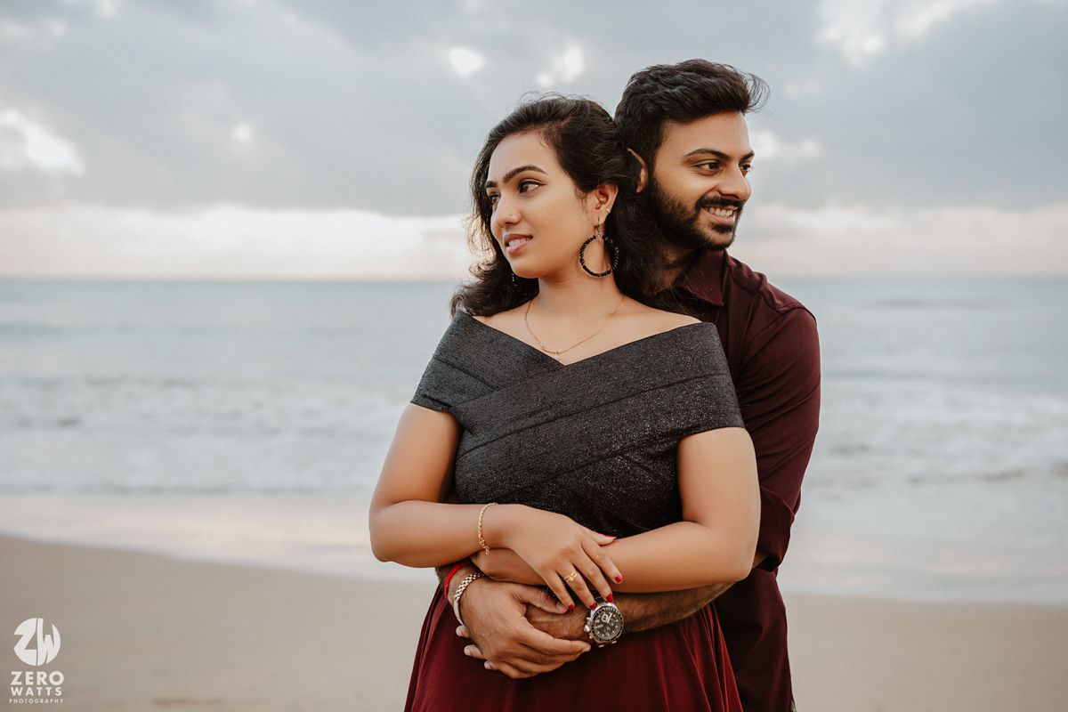 A romantic couple embraces on a beach at sunset wearing elegant formal attire in dark colors.