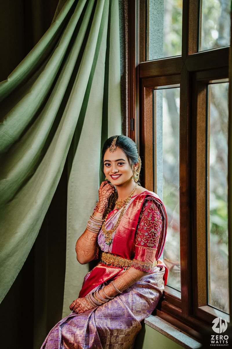 Bride in traditional silk saree and ornate jewelry stands by window with flowing green curtains in natural light.