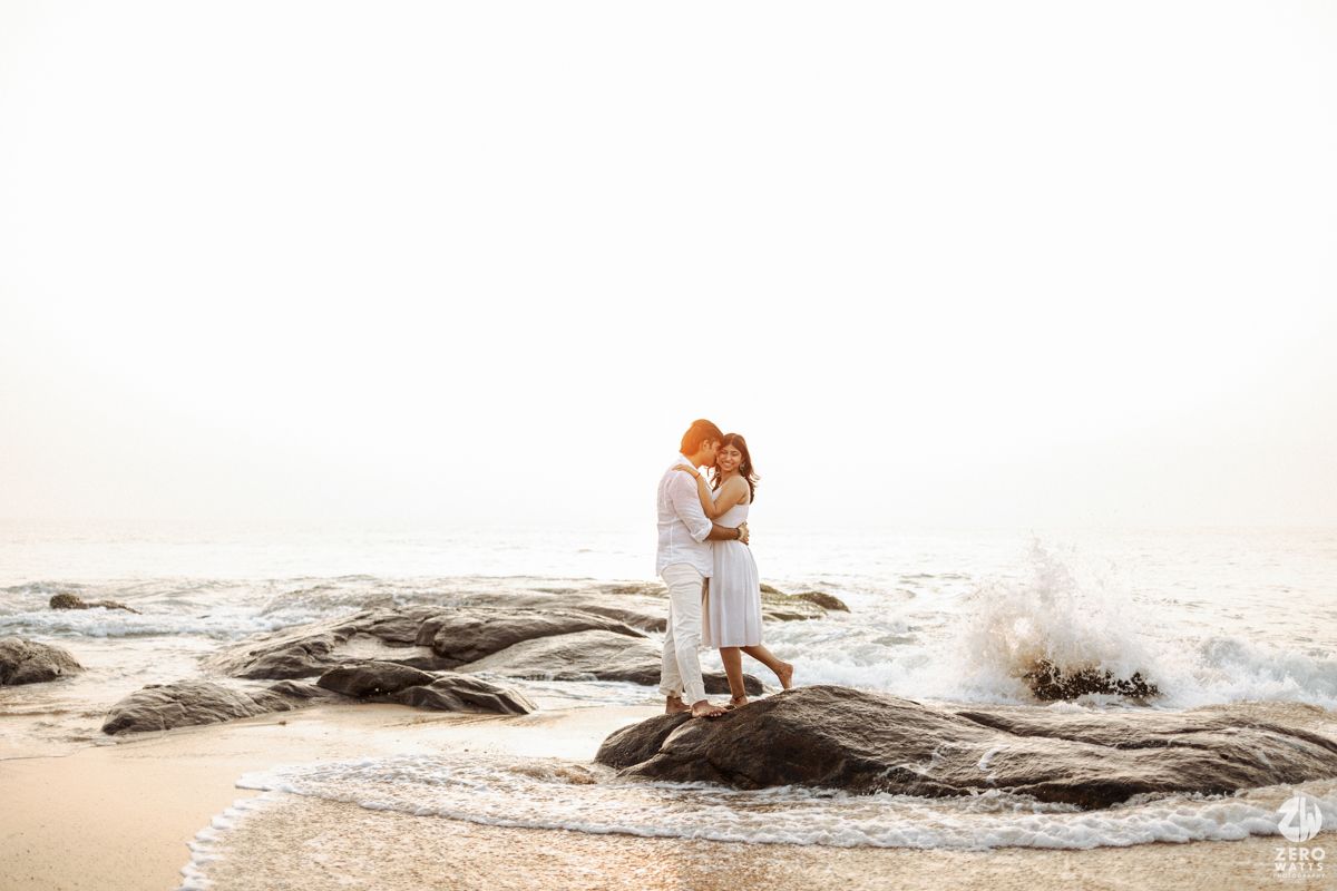 A couple in white outfits share a romantic moment on coastal rocks as ocean waves crash behind them at sunset.