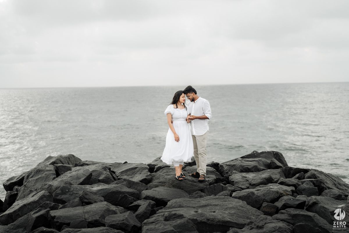 Couple in white outfits standing together on dark rocky shoreline against cloudy ocean backdrop.