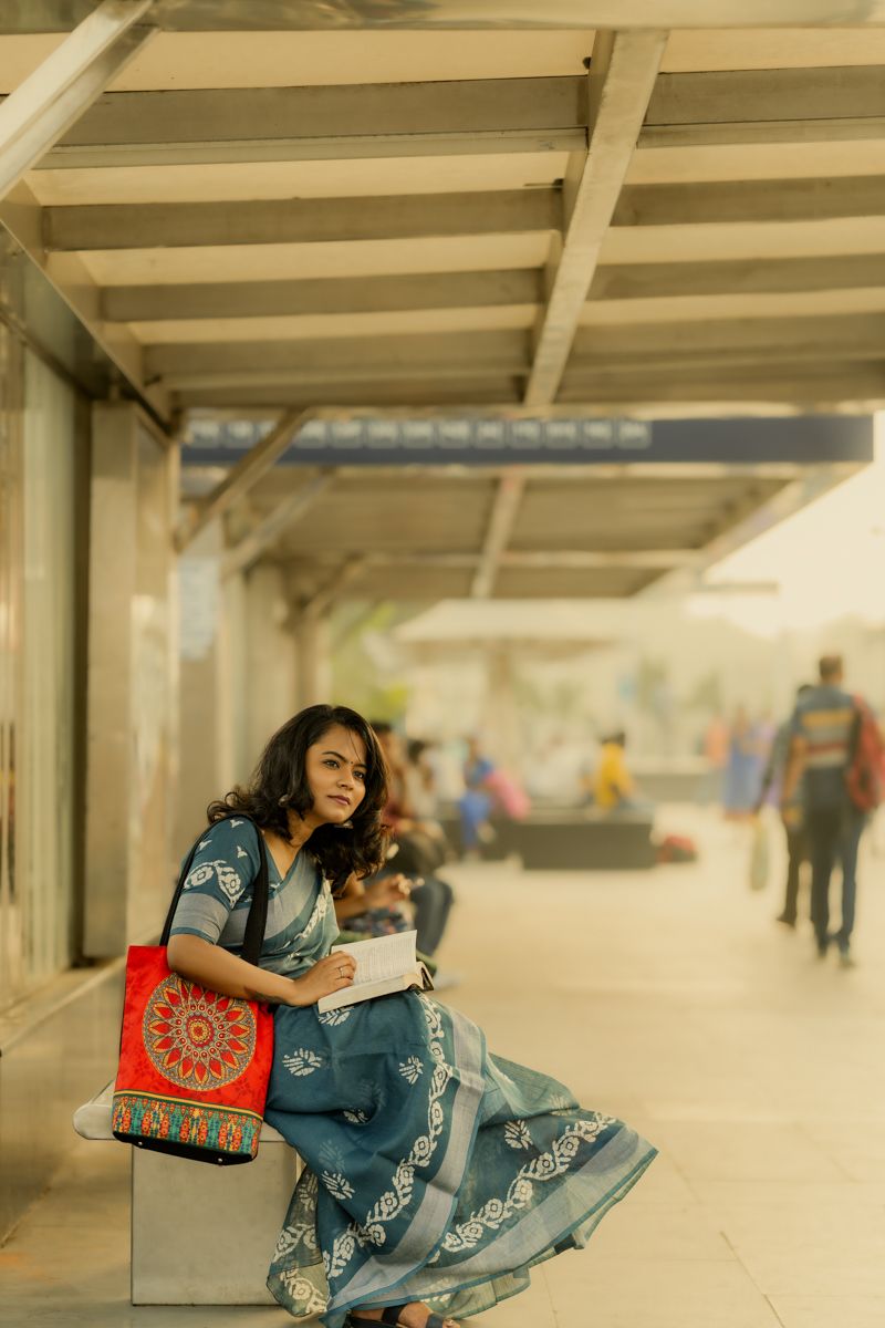 Someone in blue traditional saree holding book while sitting at train station platform with commuters in background.
