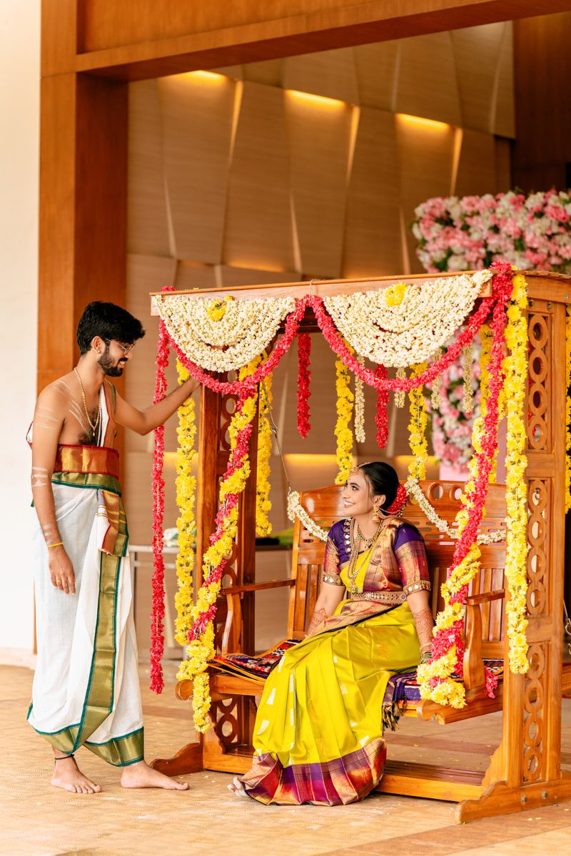Couple in traditional South Indian attire interacting near an ornate wooden swing decorated with floral garlands.