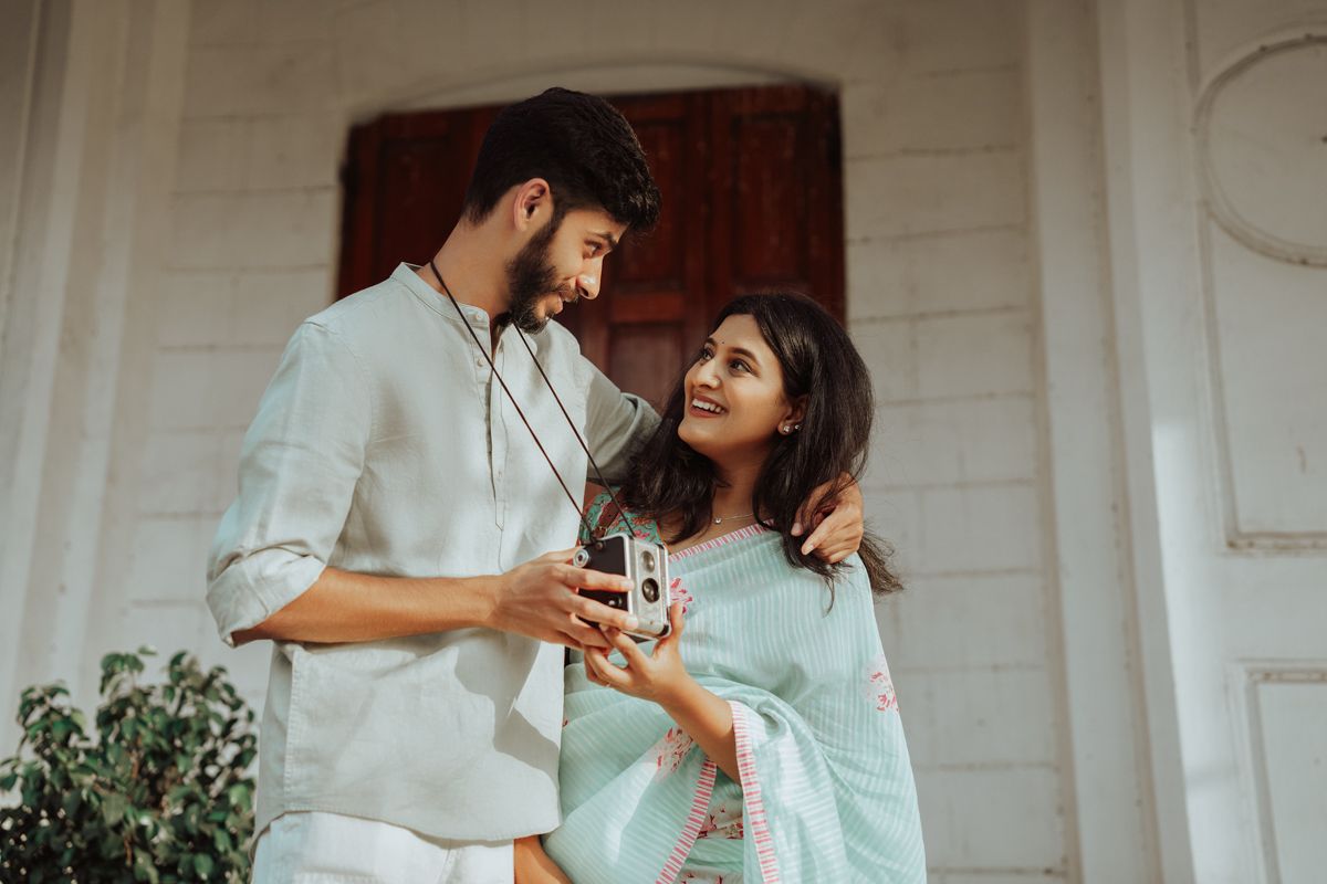 Couple in traditional Indian clothing share an intimate moment while holding tea cups on a white porch.