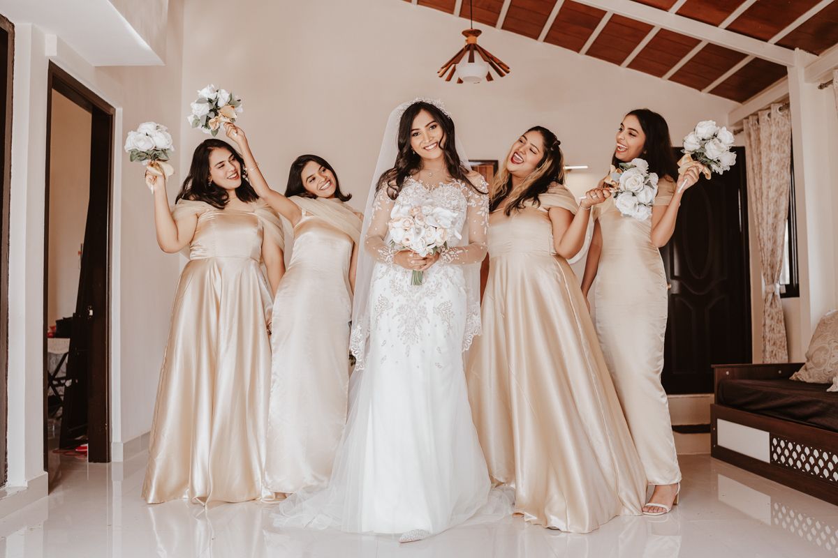 Group in elegant champagne bridesmaid dresses and white wedding gown posing joyfully in bright room.