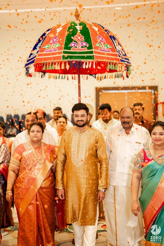 Colorful traditional Indian ceremonial umbrella decorated with tassels and designs hangs above gathering in festive setting.