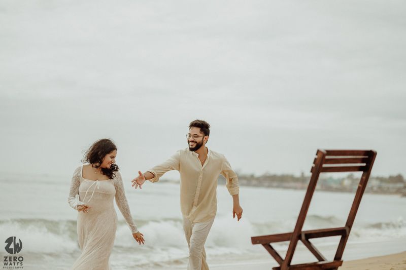 A joyful couple running and playing on a cloudy beach with wooden chair prop.