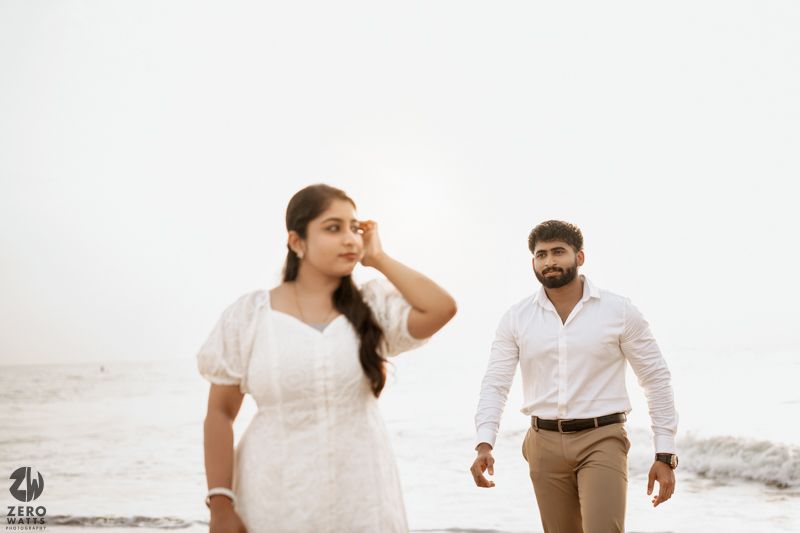 A couple in white outfits shares a playful moment while walking along a foggy beach.