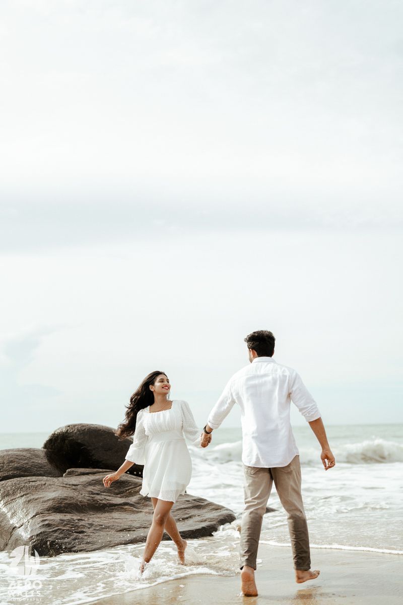 A couple walking playfully hand in hand on a beach near large rocks and ocean waves.