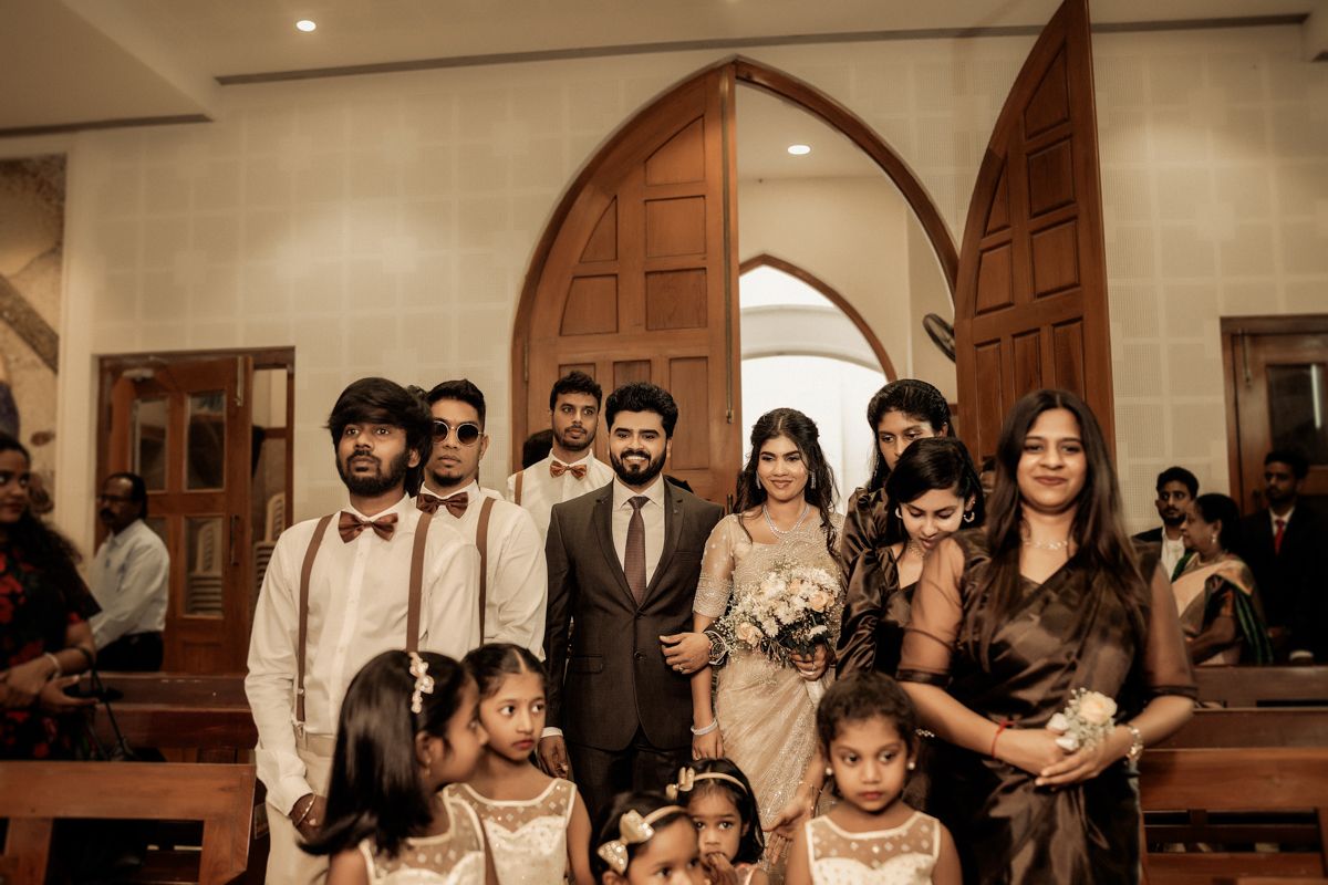 A wedding party gathers inside a church with ornate wooden arches and doors for a formal group photo.