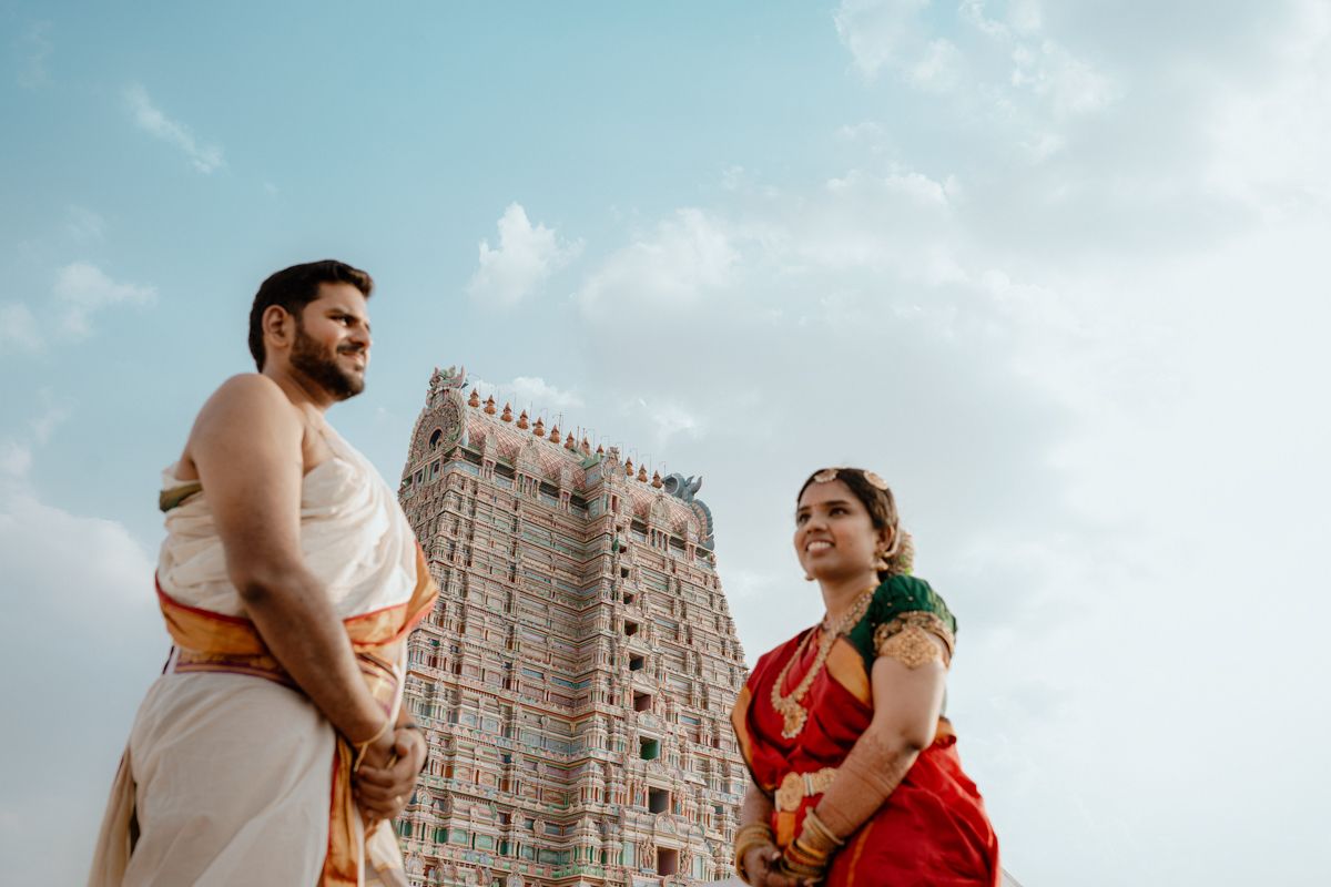 Traditional Indian couple standing in front of towering temple gopuram under cloudy sky.