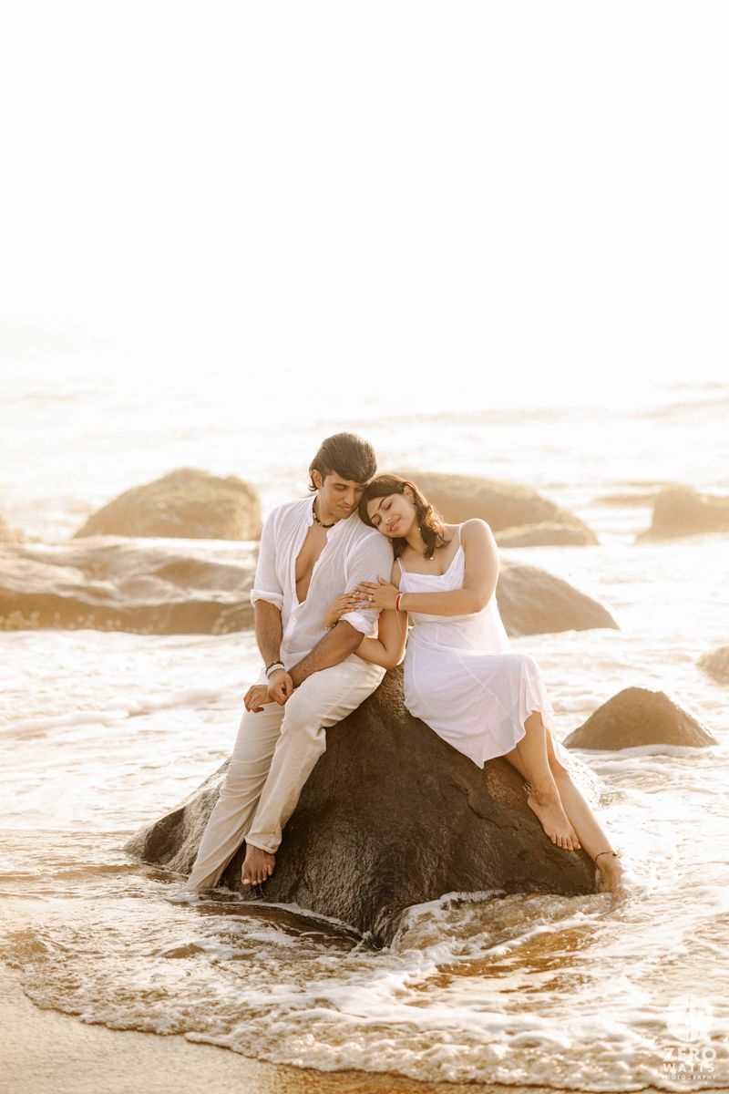 A couple in white clothing sits romantically together on a large beach rock during golden hour.