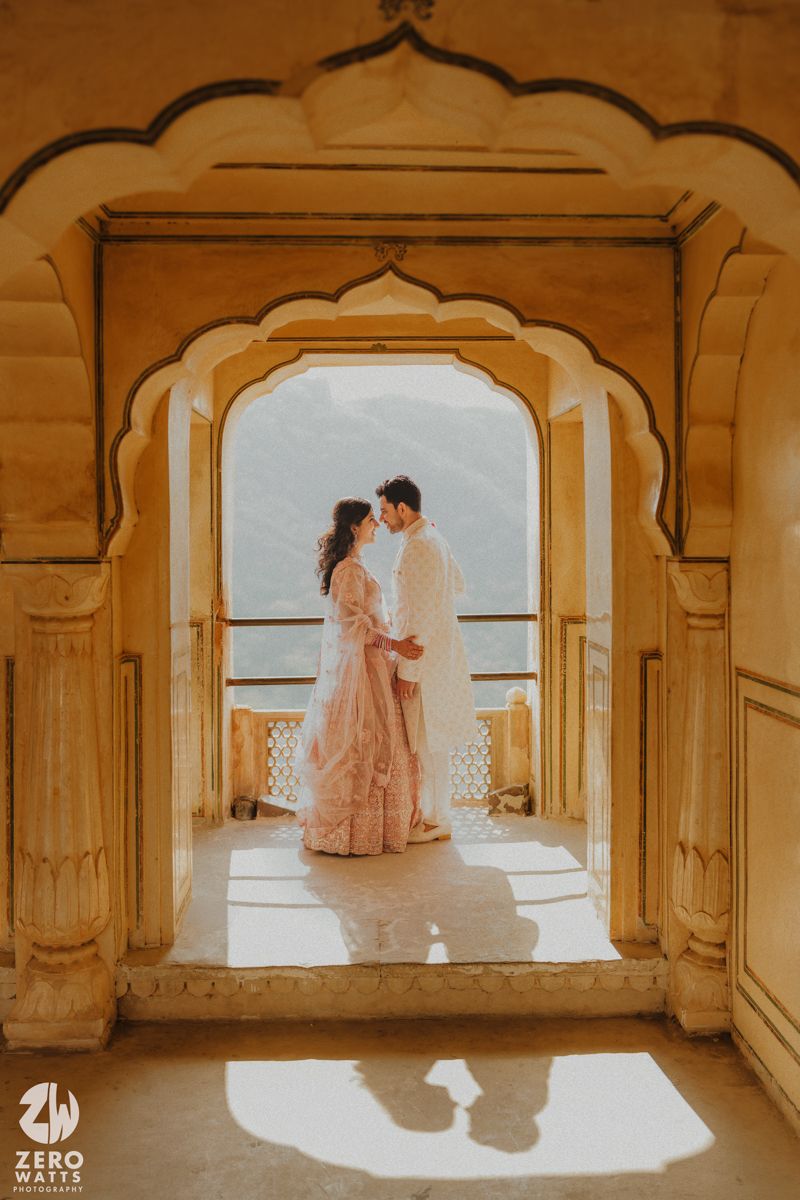 A couple stands beneath an ornate Rajasthani archway with sunlight casting shadows through traditional architecture.