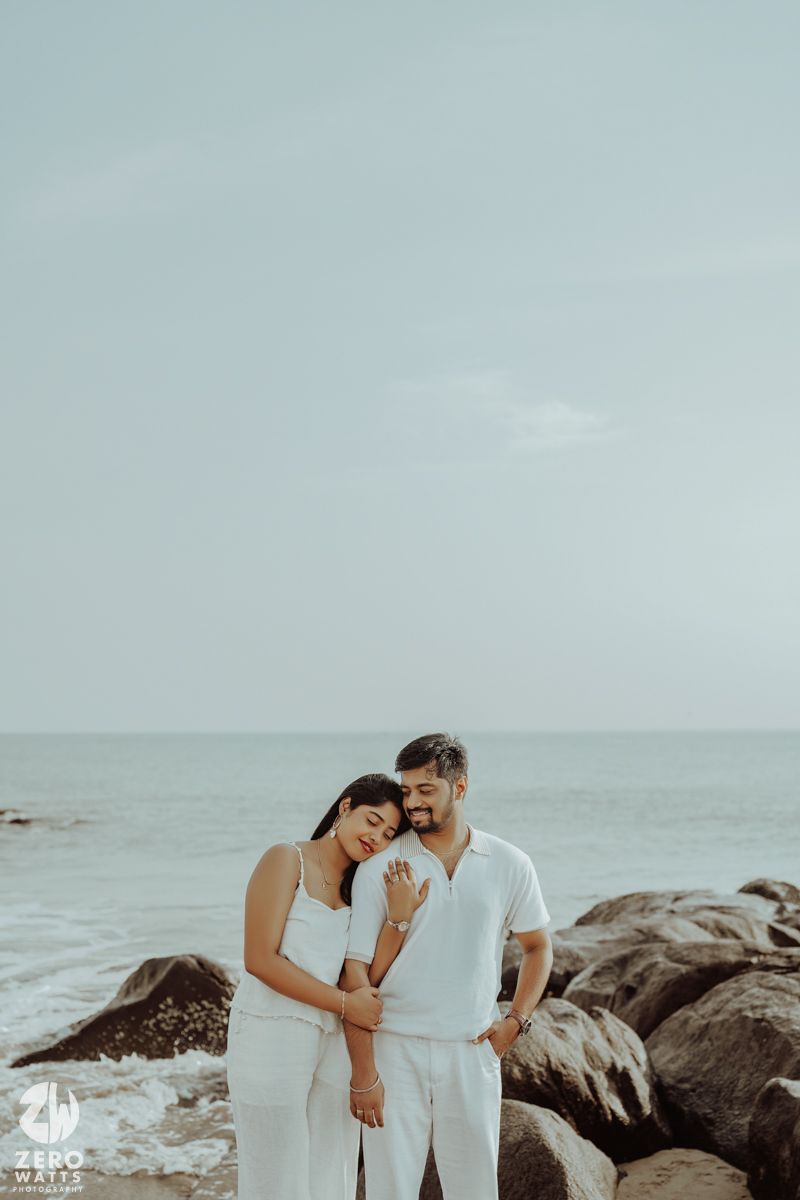 Couple in white outfits embracing on rocky coastal shoreline.
