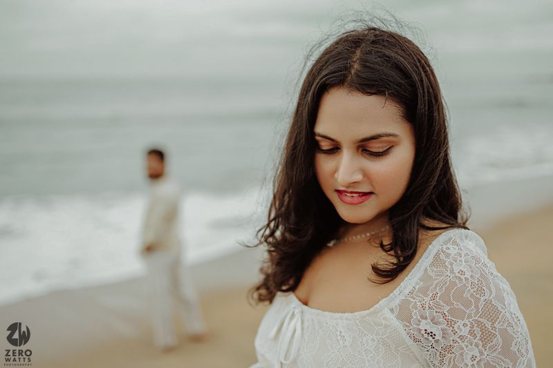 Woman in a white lace dress poses on a misty beach with soft ethereal lighting.