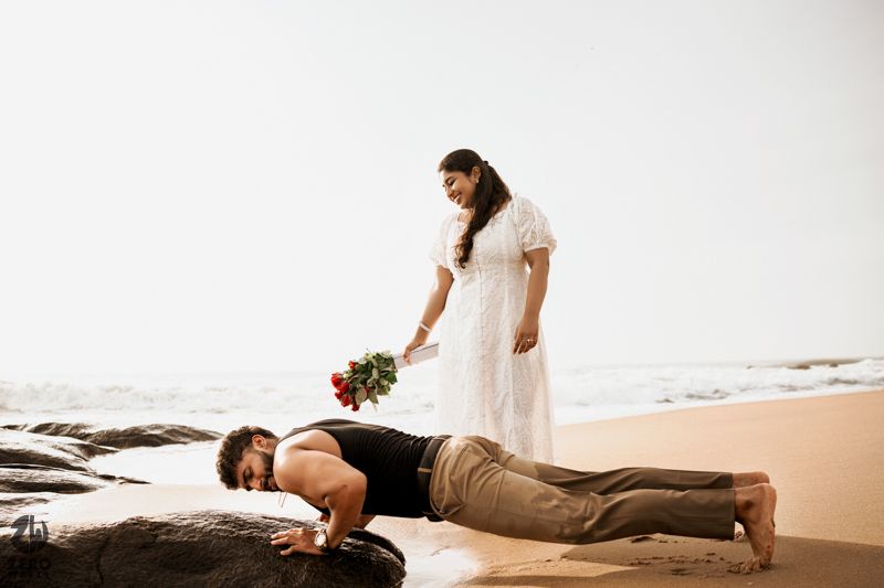 Person in white dress stands over someone doing pushups on beach rocks.