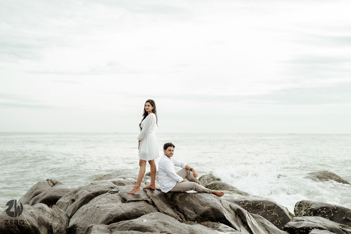 A couple in white outfits poses on rocky coastal formations with a misty ocean backdrop during an engagement photoshoot.