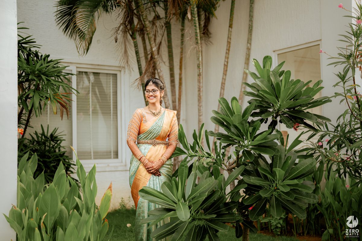 Someone in a golden dress stands among lush tropical plants and palm trees outside a building.