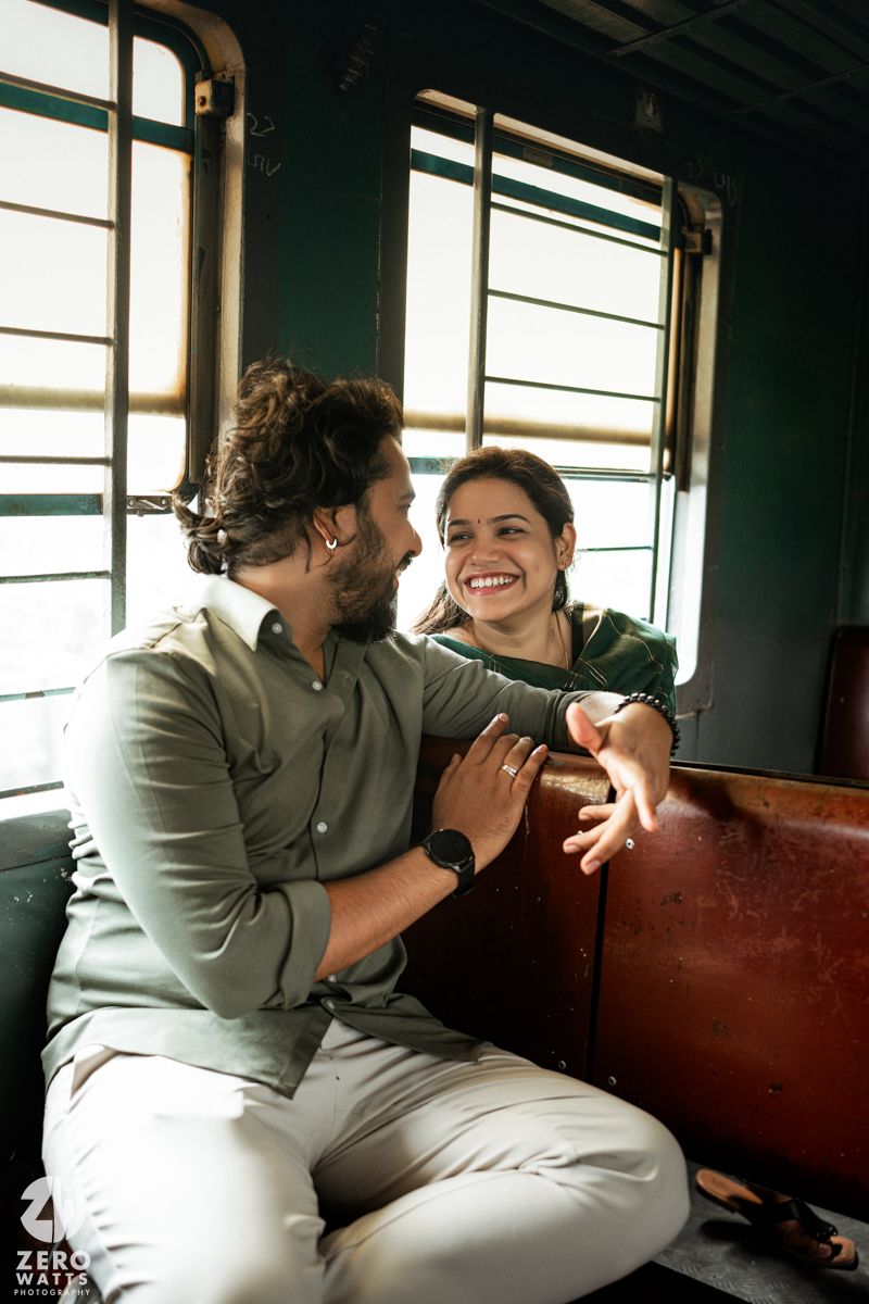 A couple shares a joyful moment together while sitting on a wooden bench in a train car with sunlit windows.