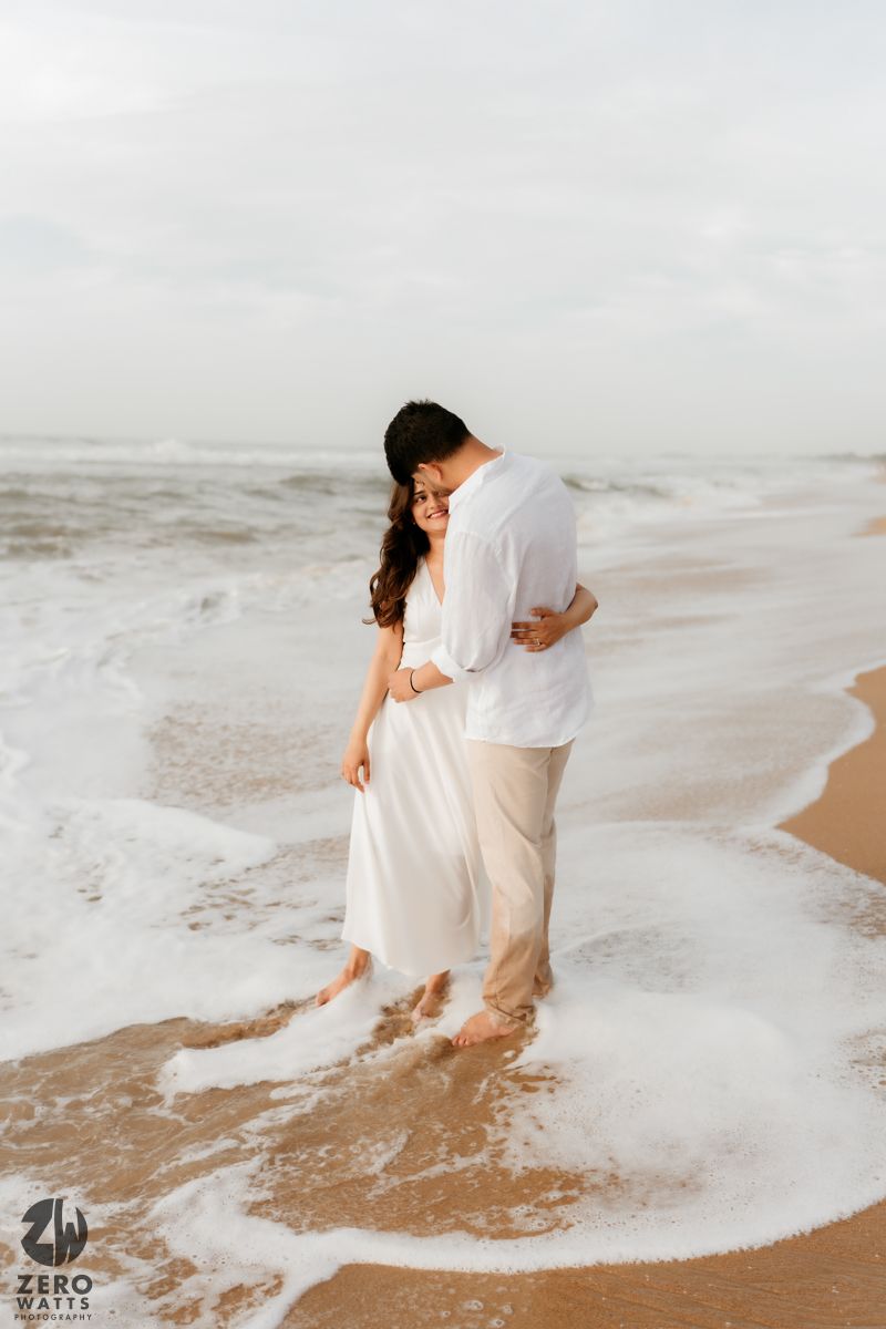 Couple in white embracing while standing in beach waves on a cloudy day.