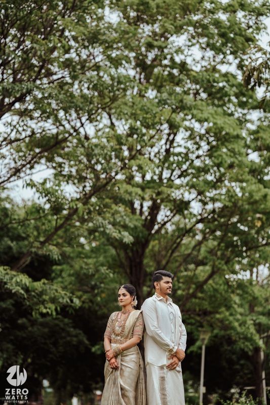 A couple in traditional wedding attire poses outdoors under leafy green trees in a romantic garden setting.