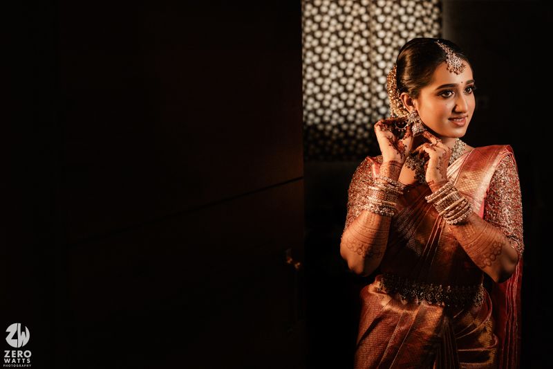 Traditional Indian bride in coral silk saree adorned with jewelry gestures expressively during wedding celebrations.