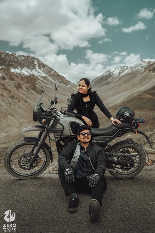 Adventure motorcyclists pose with their bike against dramatic snow-capped mountains and turquoise sky in a remote location.