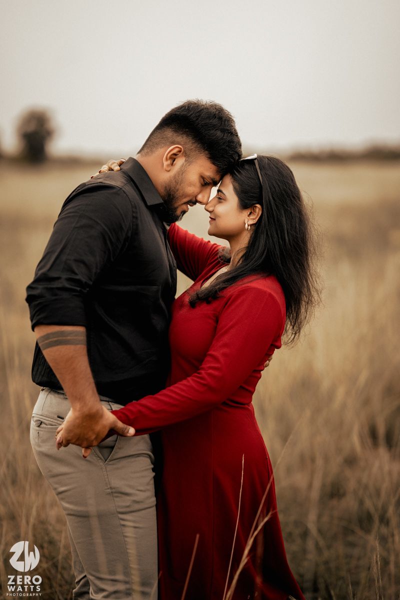 A romantic couple in red and black outfits share an intimate moment in a rustic field setting.