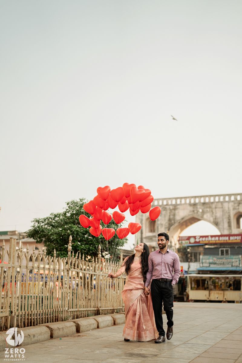 A romantic couple walks along a promenade holding red heart balloons against architectural backdrop.