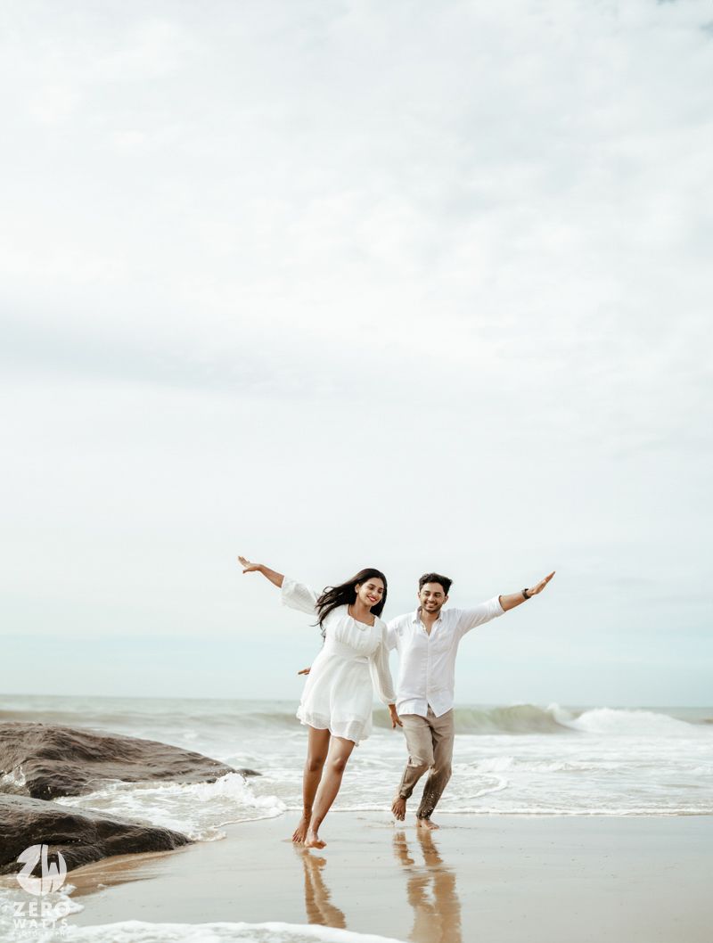 A joyful couple jumps with arms spread wide on a scenic beach with waves in the background.
